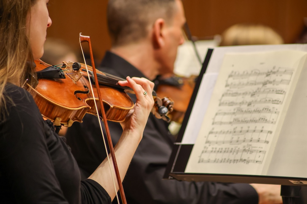 Close-up of violinists playing in an orchestra, with one musician’s hand and sheet music in clear focus.