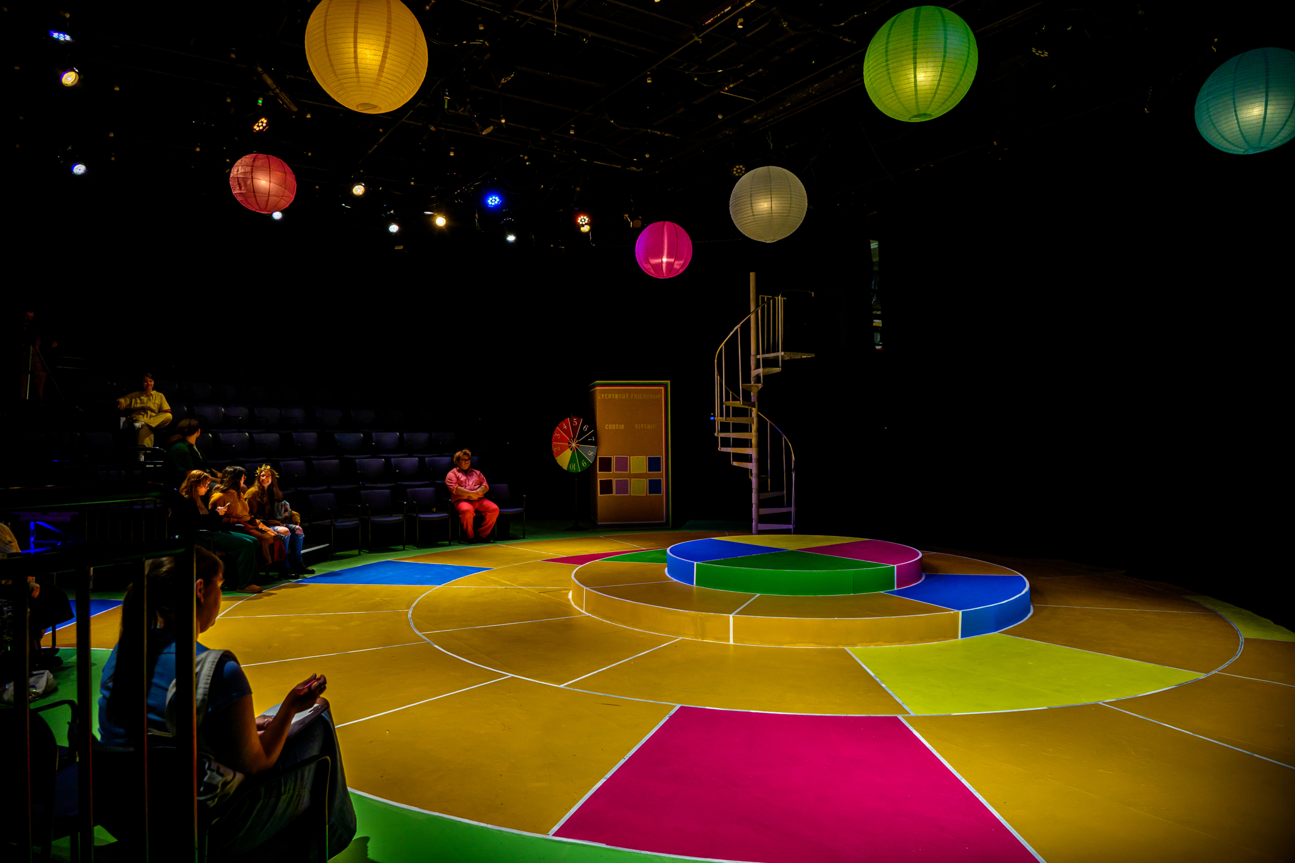 Photo by Chuck Steenburgh A round, multicolored stage with tiered platforms is set under hanging lanterns, with a small audience seated in a darkened theater.