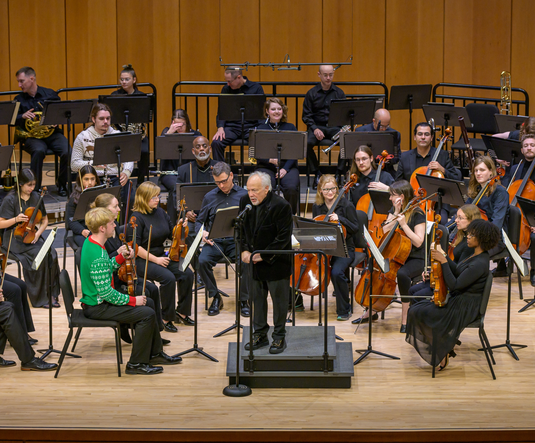 A conductor stands at a podium in front of an orchestra on stage, with musicians holding various instruments and seated in rows.