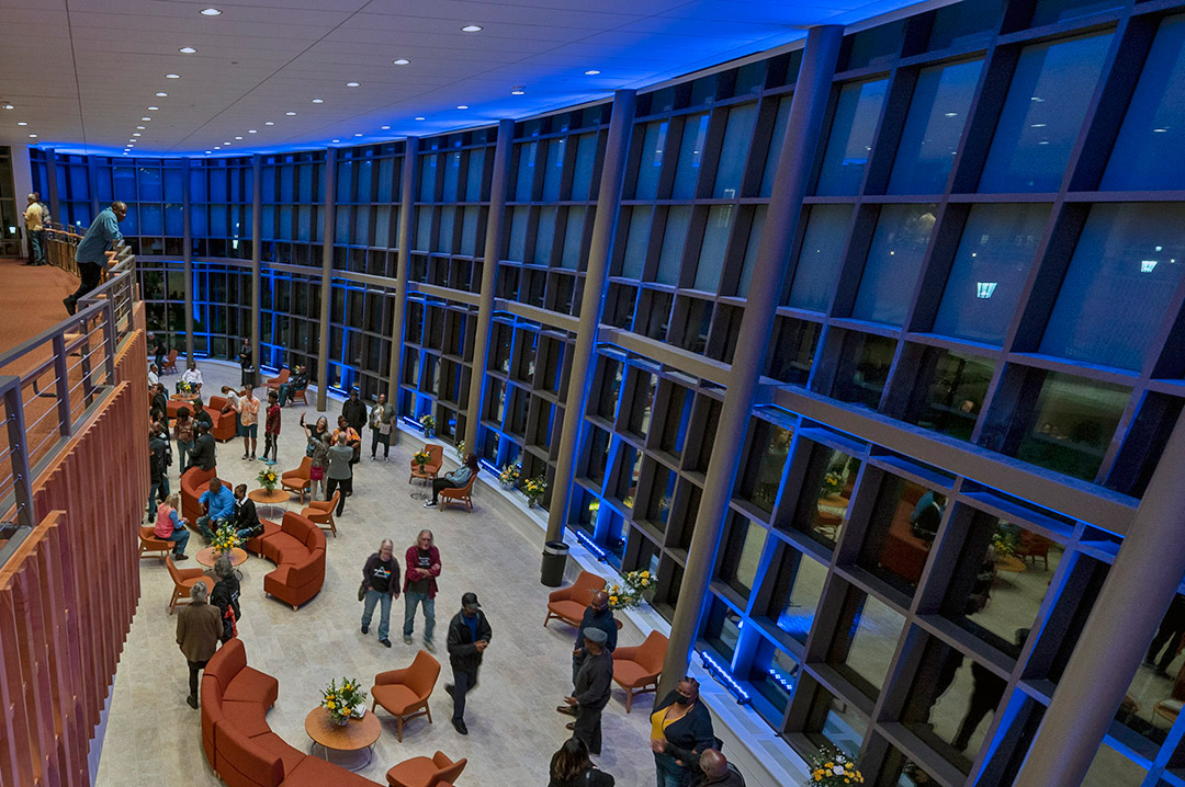 DodgePAC-Lobby1 People gather and converse in a spacious, modern lobby with large glass windows and blue accent lighting in the evening.