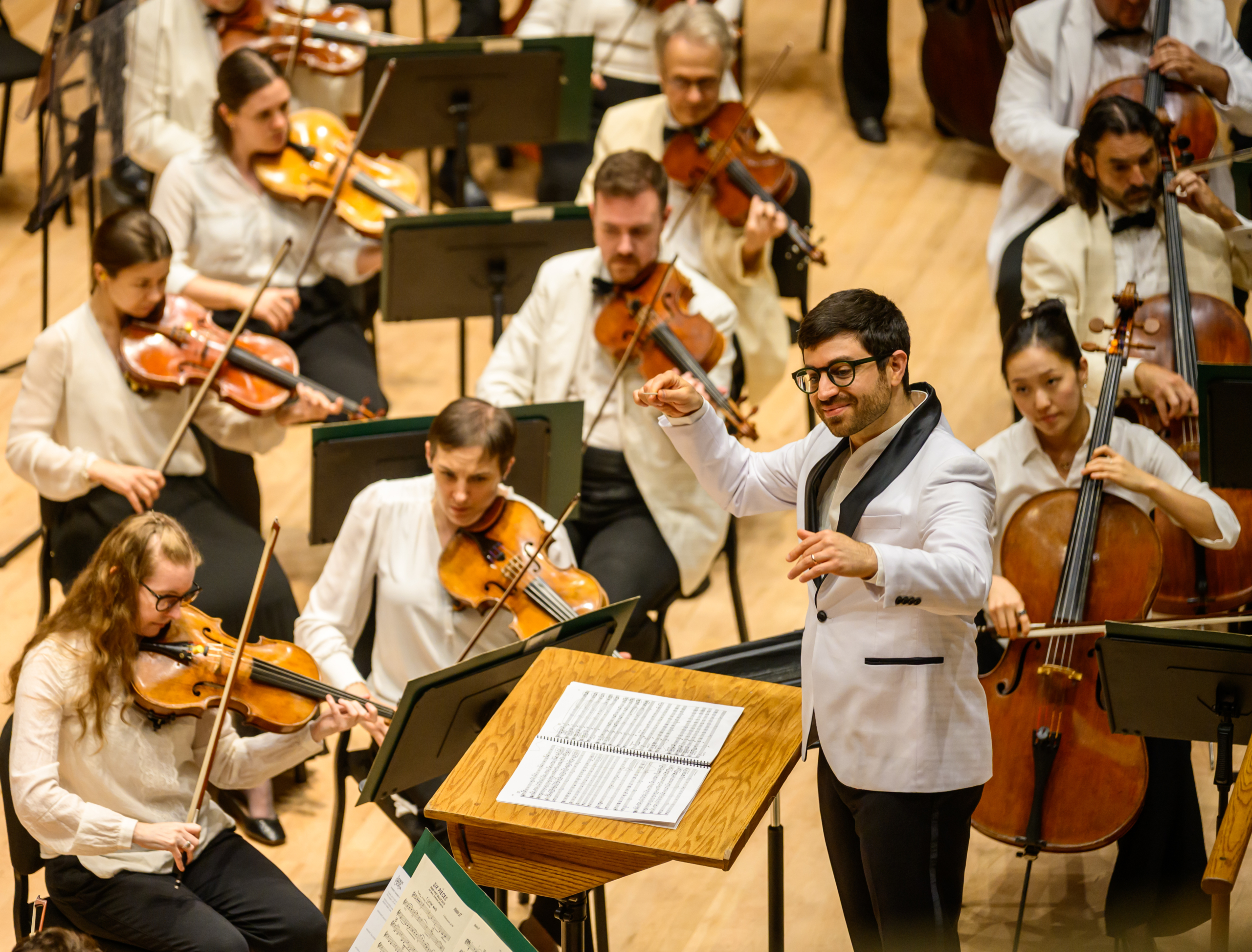 A conductor leads an orchestra of string musicians on stage, all wearing white jackets and performing with sheet music on stands.