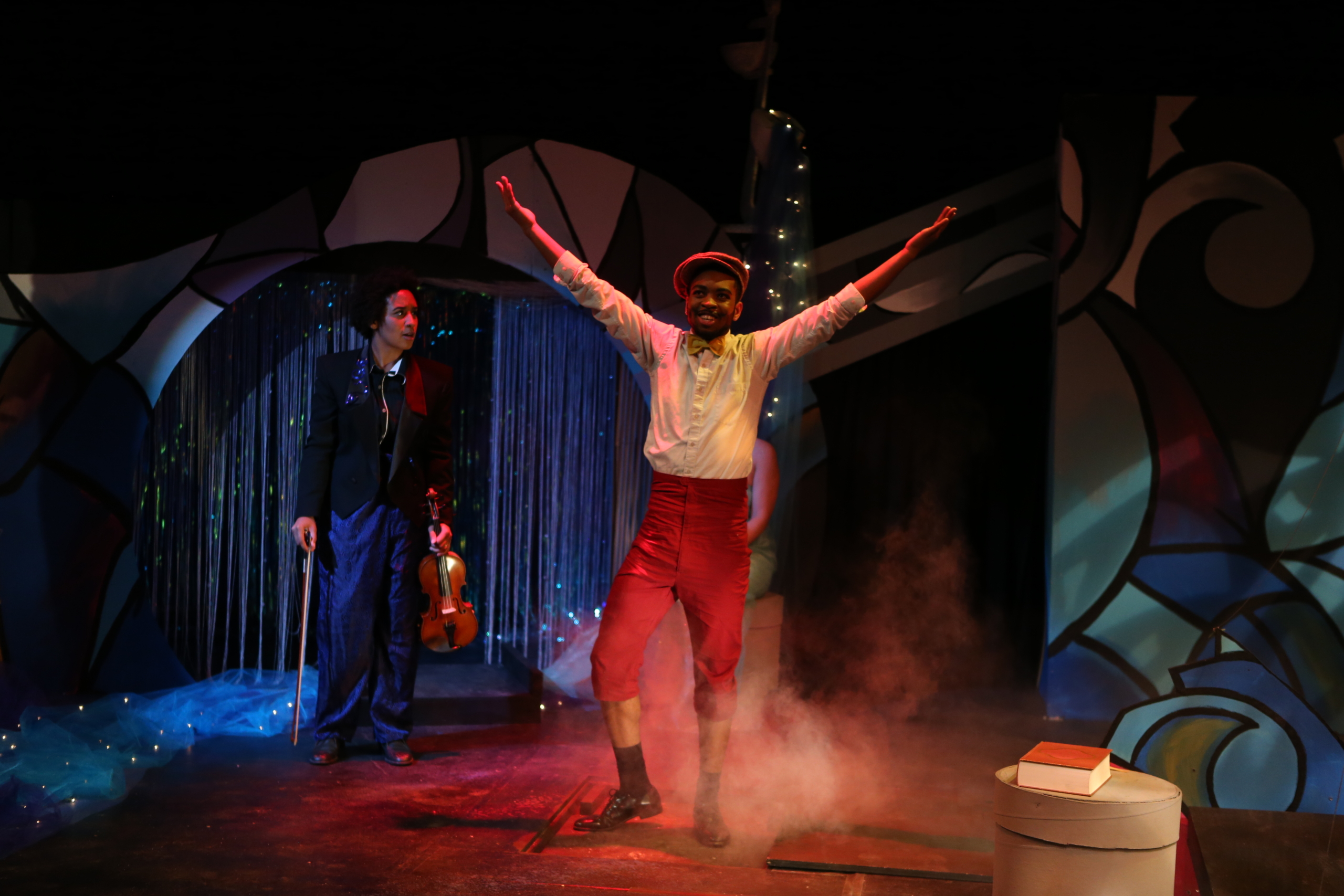 EA9Q9357 A performer in a white shirt and red pants stands with arms raised center stage, while another person with a violin stands behind him under theatrical lighting and stage decorations.