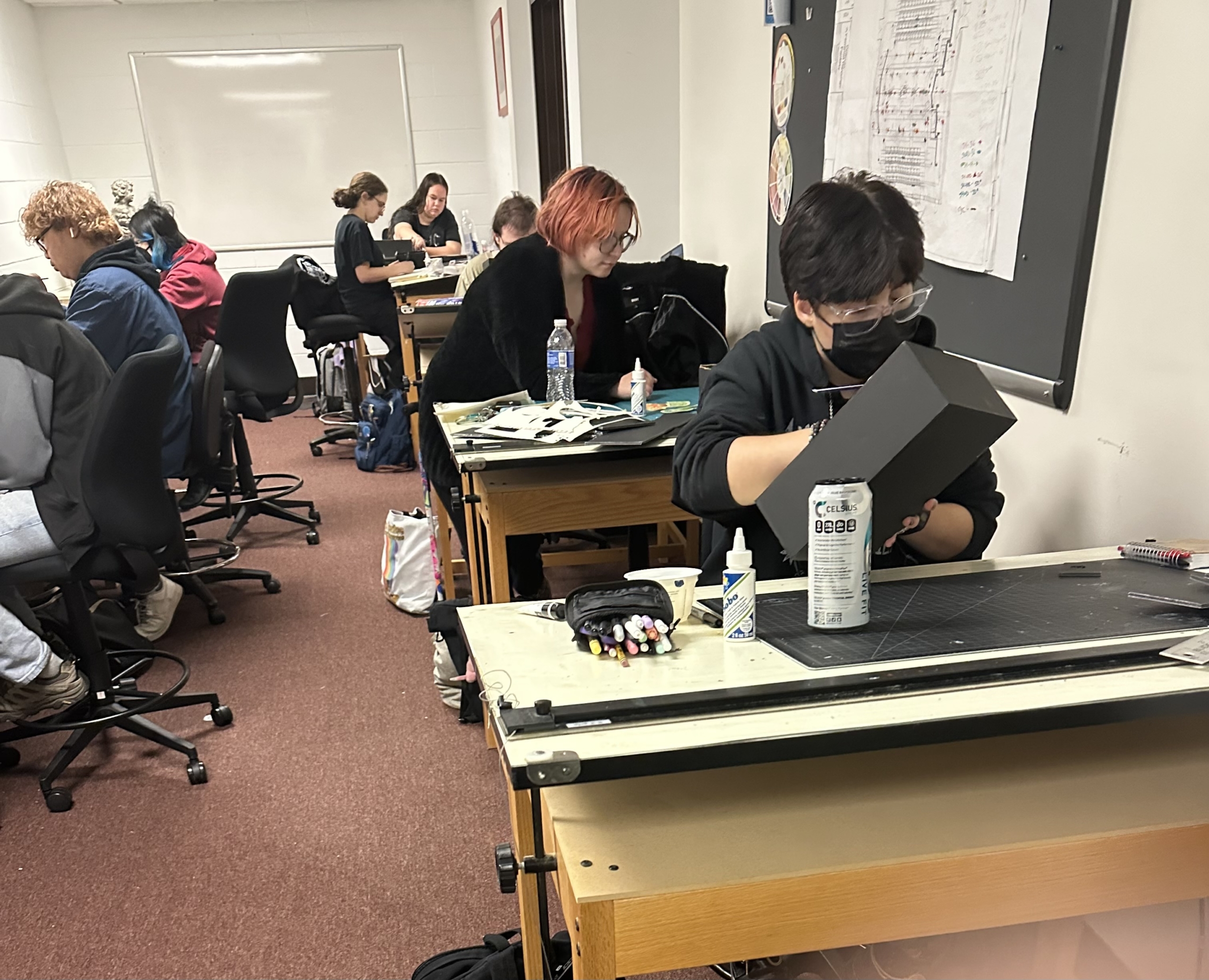 Students work on art projects at desks in a classroom, using various supplies like markers, glue, and cutting tools. Some appear focused on their tasks, while others interact or observe.