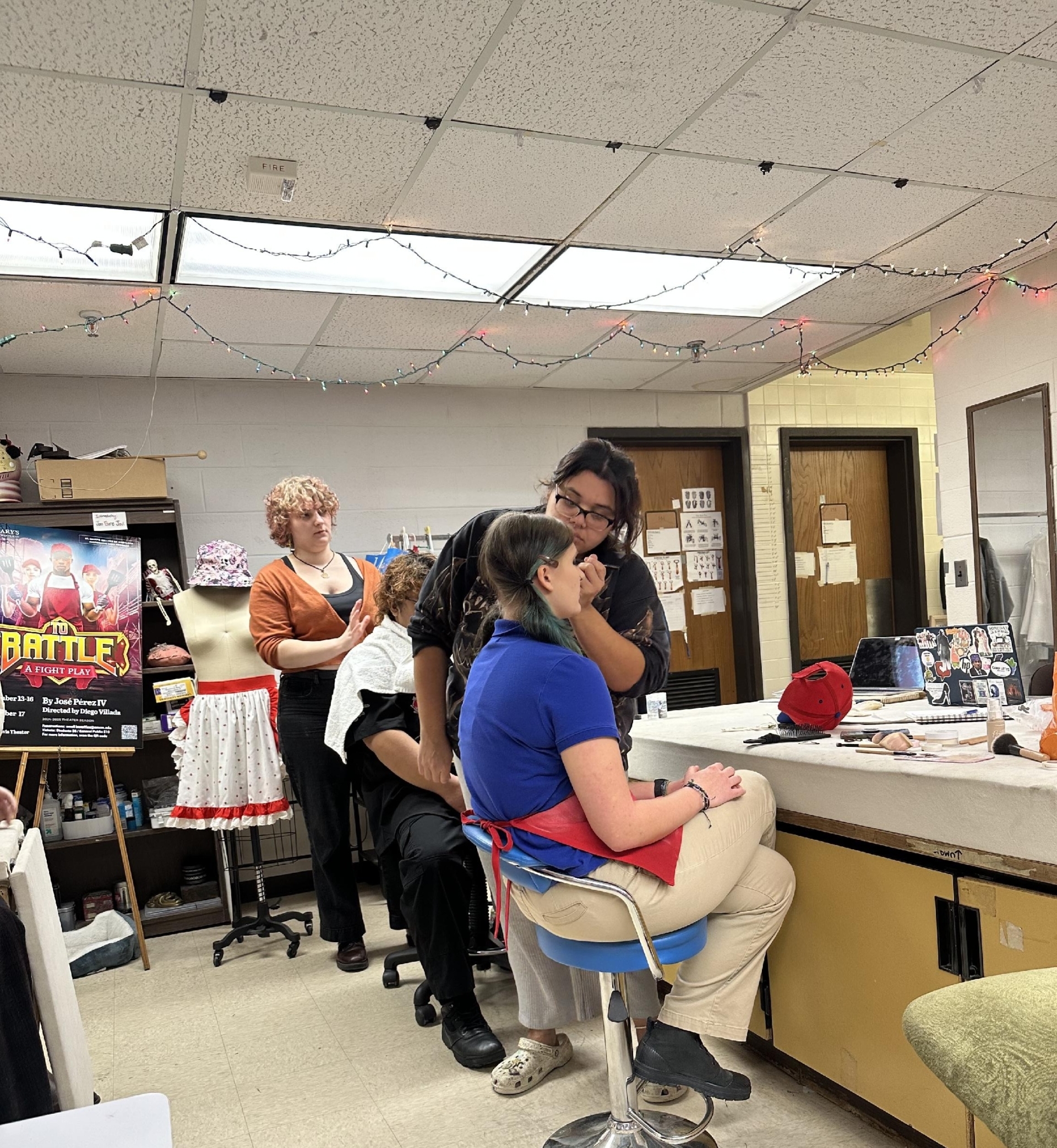 Four people are in a backstage room; one person applies makeup to another seated person, while two others stand nearby. Costumes and a "BATTLE" poster are visible in the background.