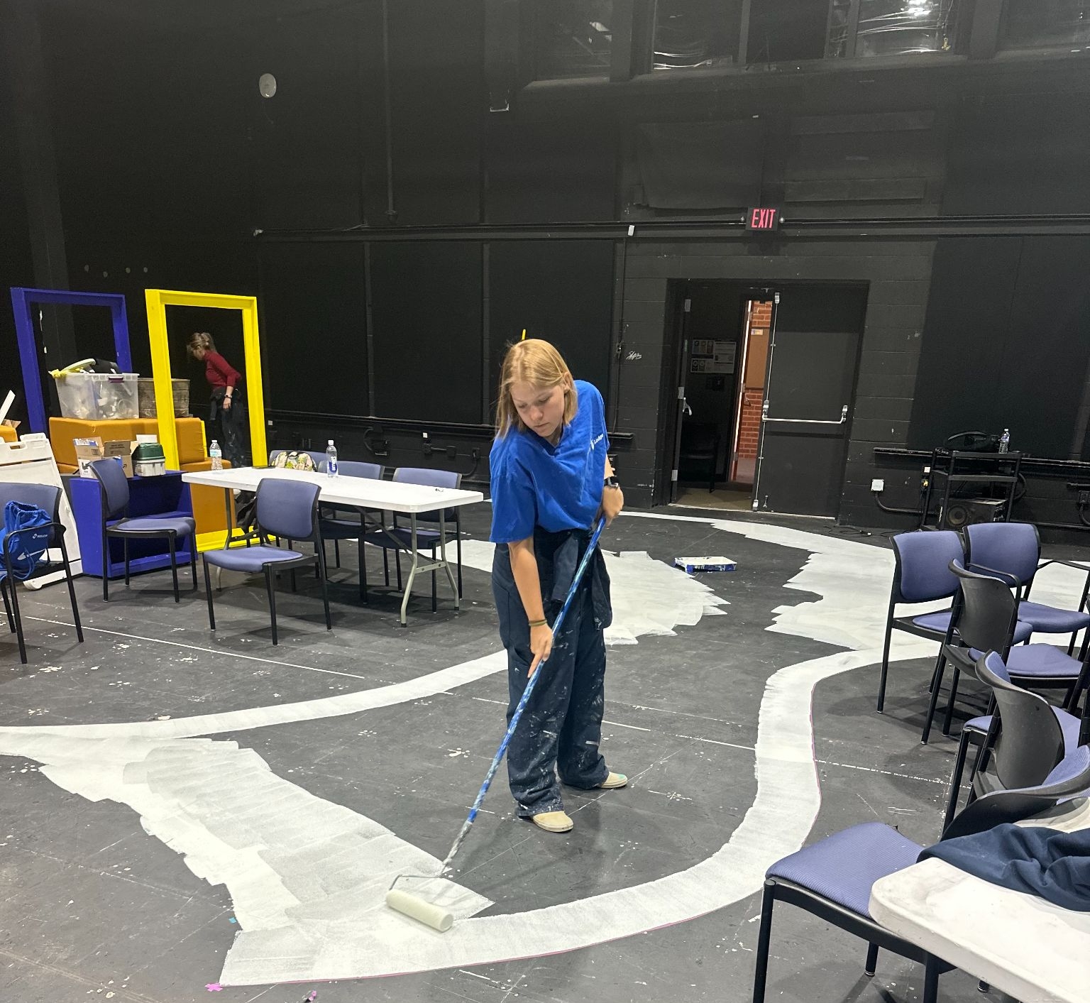 A person uses a paint roller to apply white paint to a large, irregular area on the floor of a room filled with scattered chairs and tables.
