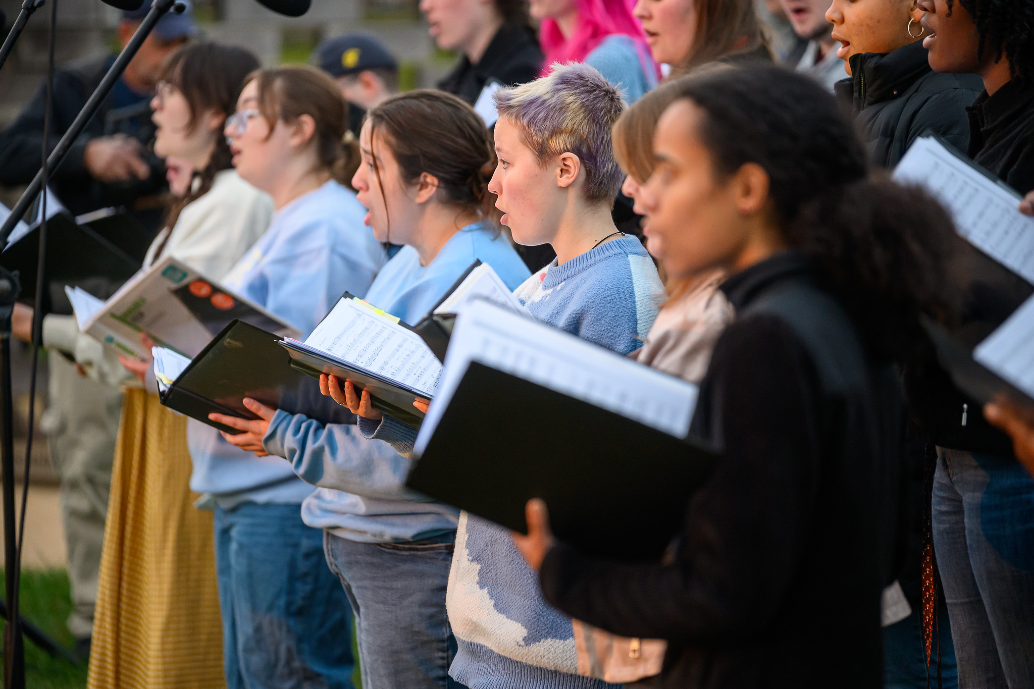 A group of people stand closely together outdoors, holding sheet music and singing in a choir performance.