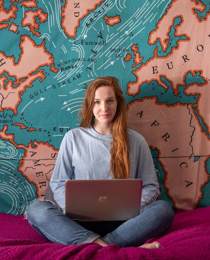 31349034747_3be9edfe45_o A person with long red hair sits cross-legged on a bed using a laptop, with a large world map tapestry hanging on the wall behind them.