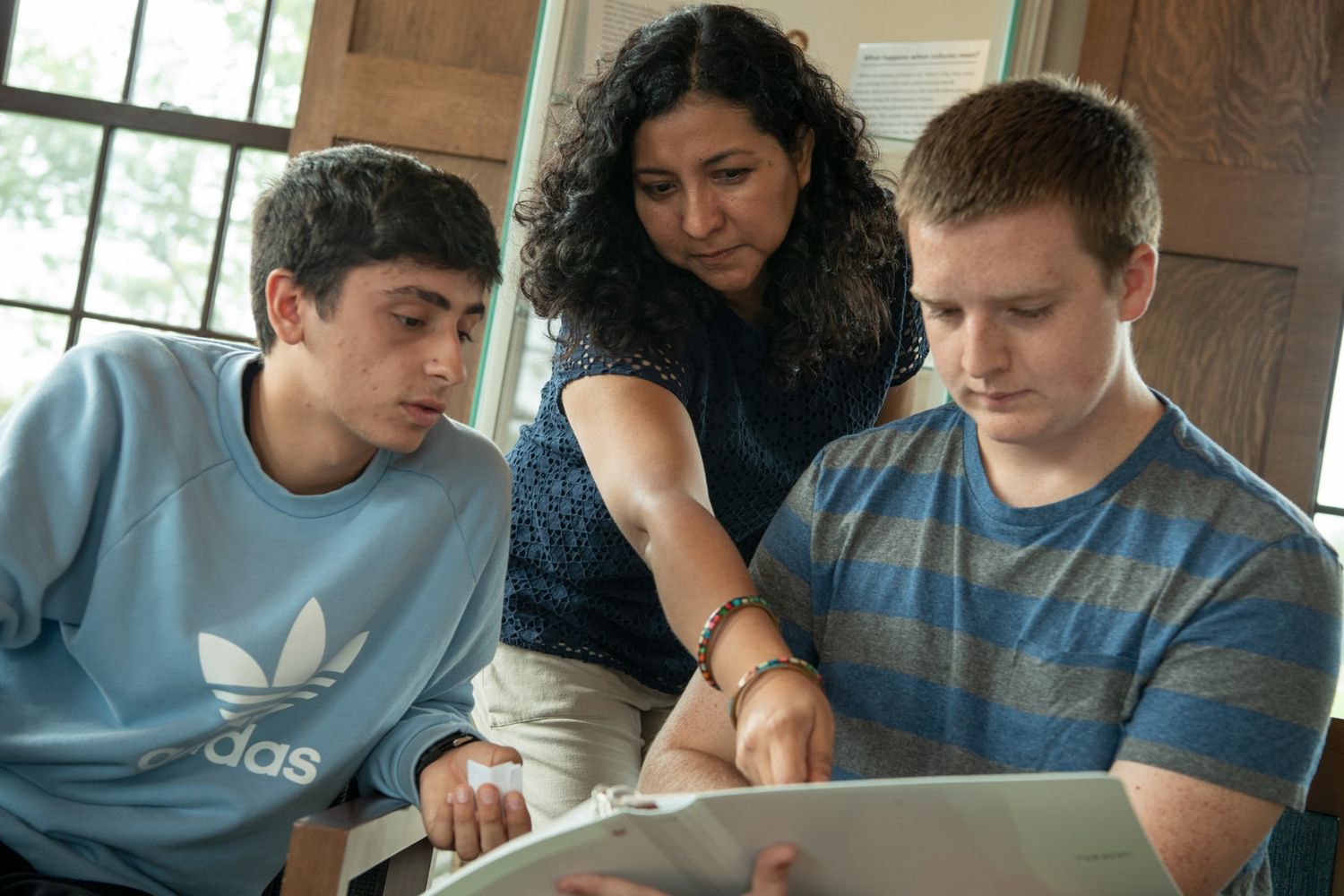 A woman points at a binder while two young men look on attentively, sitting indoors near a window.