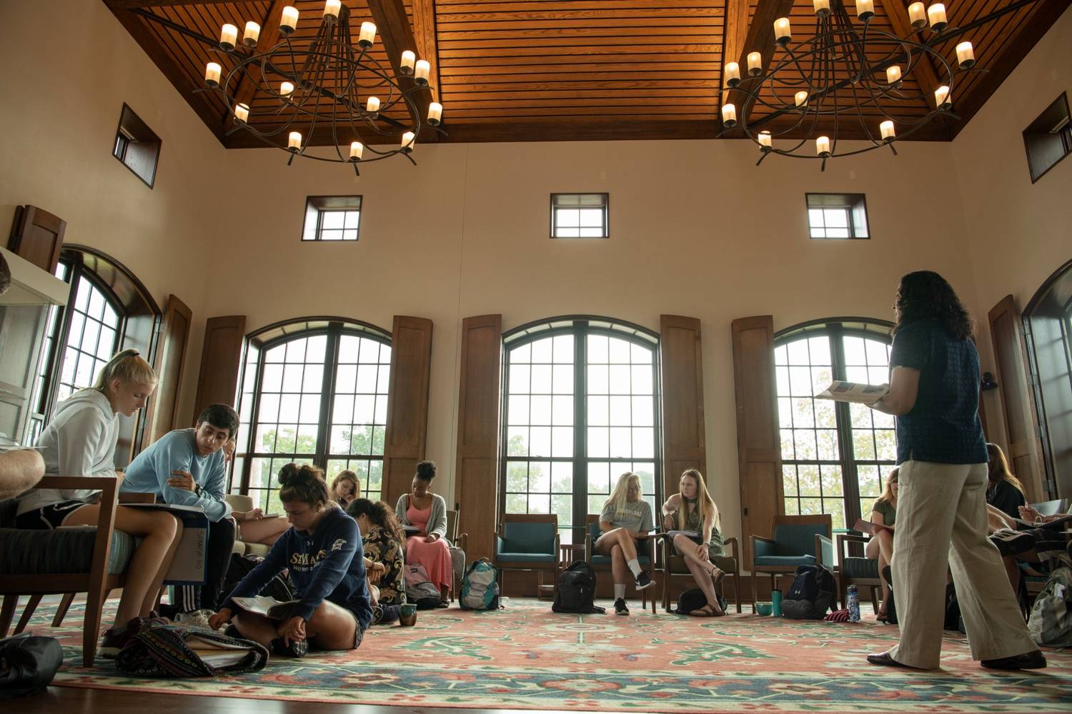 Students sit in a spacious, well-lit room with large windows, listening to an instructor standing at the front of the class.