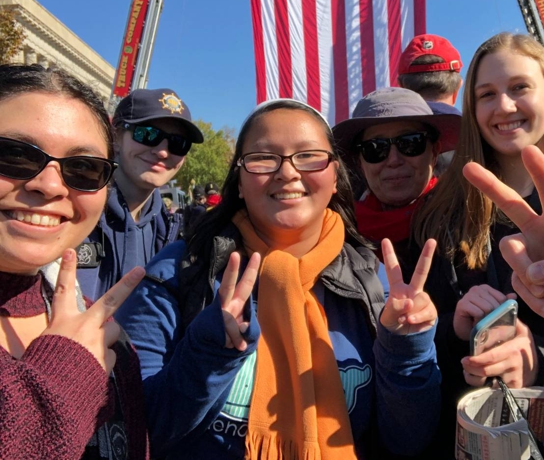 50888884652_f400bc6ca1_o A group of people smiles and holds up peace signs in front of a large hanging American flag at an outdoor event.