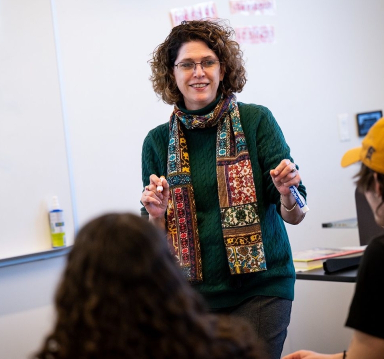 52768894589_f2ef119bcc_o A woman wearing glasses and a patterned scarf stands and speaks in front of a whiteboard to students in a classroom.
