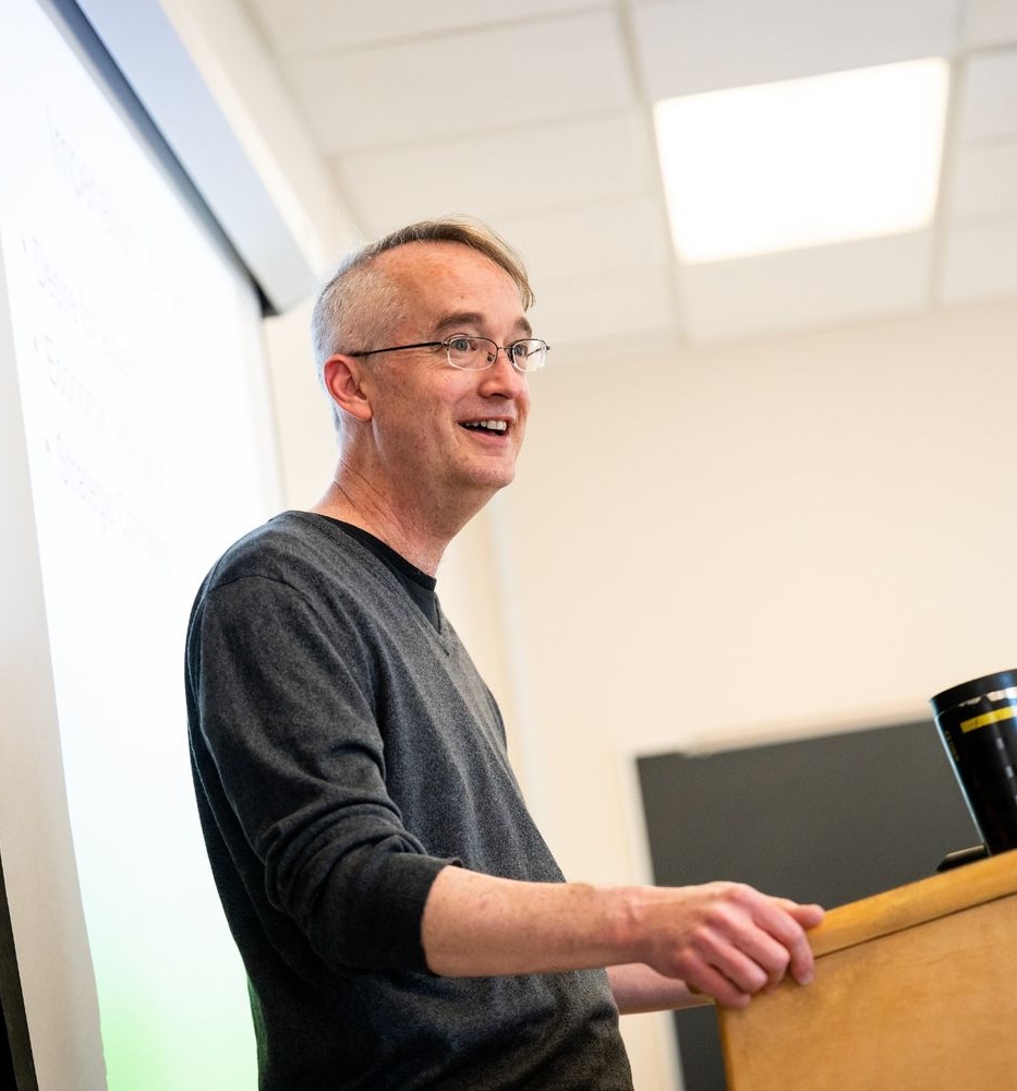A man with glasses stands at a wooden lectern, speaking in a classroom with a chalkboard and projection screen behind him.