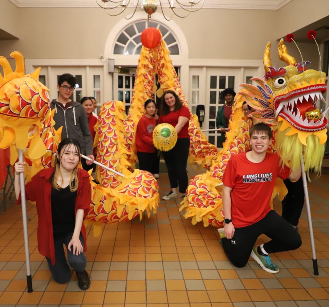 53529347396_8a13615186_o A group of people pose indoors holding a yellow and orange dragon costume, with a tiled floor and arched windows in the background.