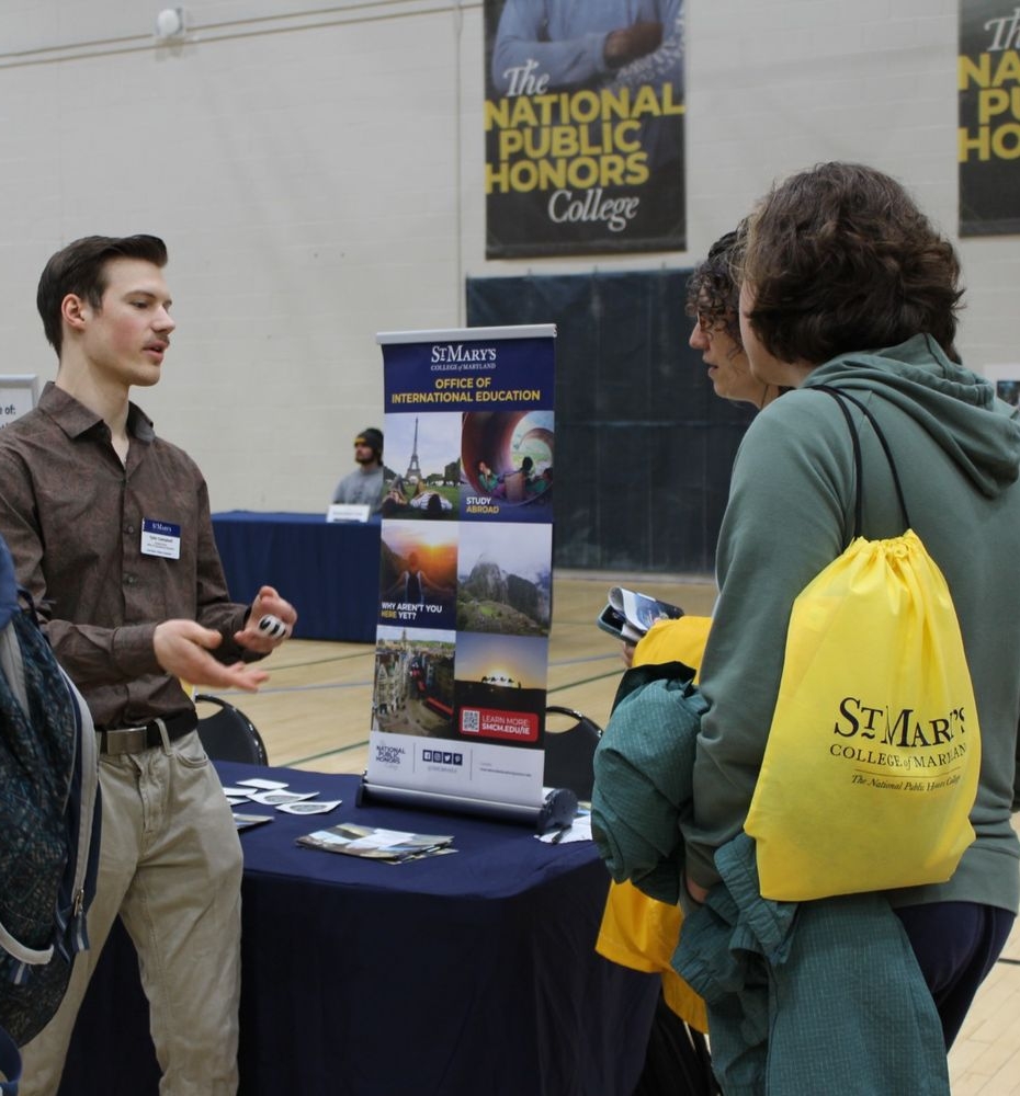 54443846204_1c2133d4cf_o A man stands at an information booth talking to three people in a gymnasium; a banner and promotional materials for St. Mary’s College of Maryland are visible.