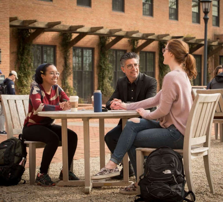 2131-1592 Three people sit and talk at an outdoor table, with backpacks and drinks visible, in a courtyard area near a brick building.