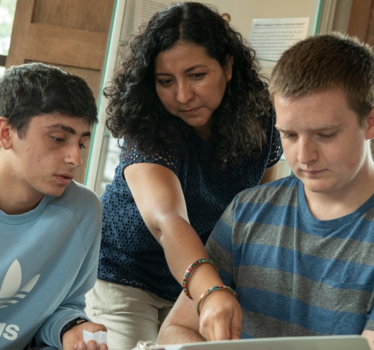 45375197605_fc6b096087_o A woman points at a laptop screen while two young men look on, appearing focused and engaged in a discussion or lesson.