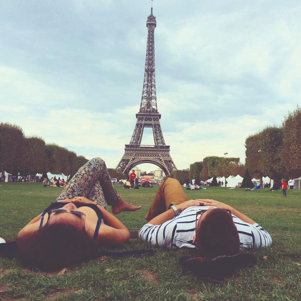 LAND, Luke – Paris Nap Two people lie on the grass facing the Eiffel Tower in Paris, with trees and other visitors in the background under a cloudy sky.