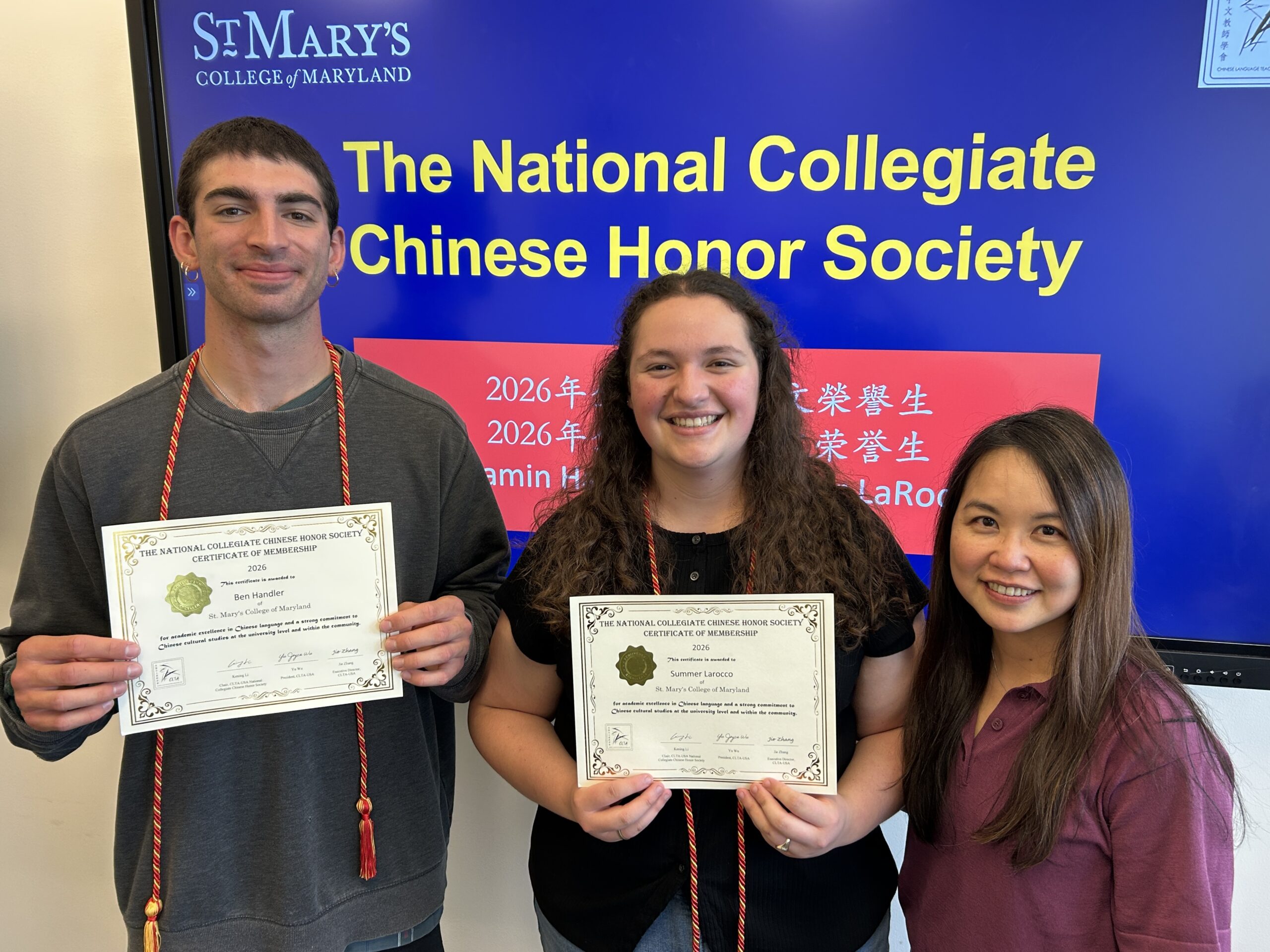 Two students hold certificates while standing next to a woman in front of a screen displaying "The National Collegiate Chinese Honor Society" at St. Mary's College of Maryland.