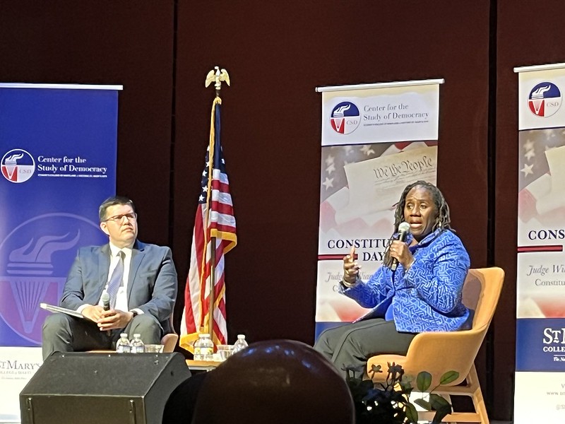 Two people are seated on stage having a discussion, with an American flag and banners for the Center for the Study of Democracy in the background.