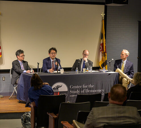 Four men sit at a panel discussion table in front of an audience, with banners, a projector screen, and flags behind them at the Center for the Study of Democracy event.