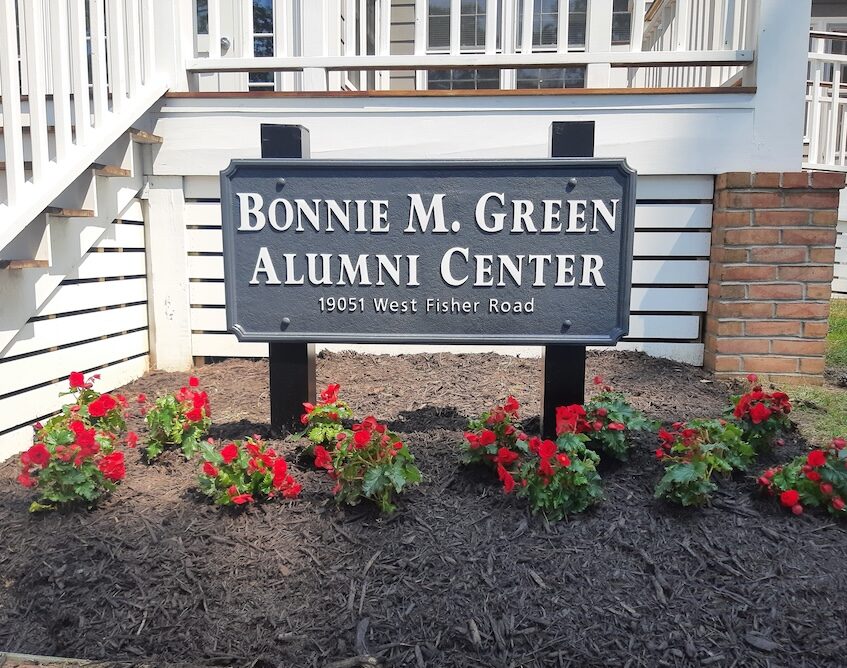 A sign reads "Bonnie M. Green Alumni Center, 19051 West Fisher Road" in front of a building, surrounded by red flowers and mulch.