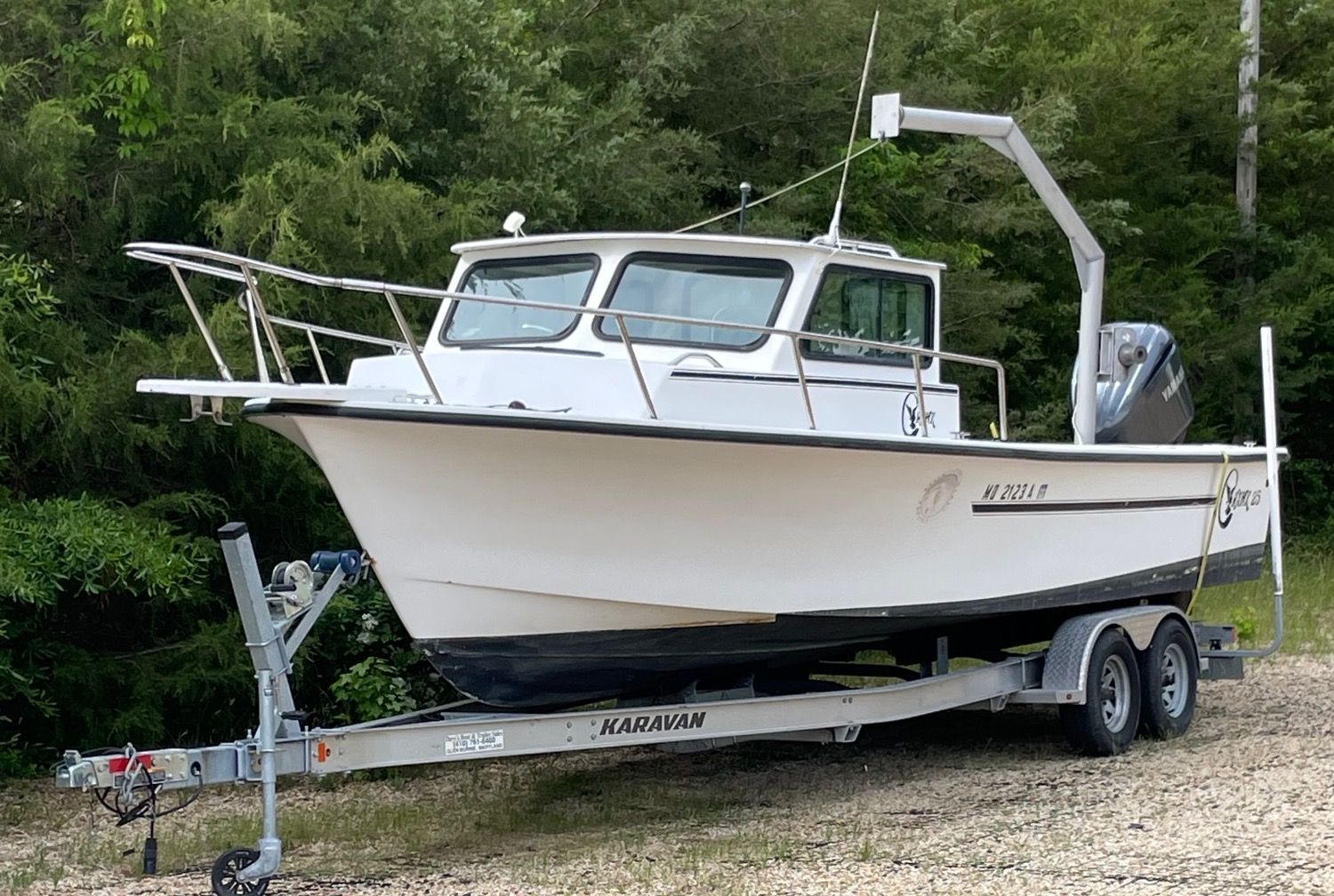 White motorboat on a dual-axle trailer parked on gravel, surrounded by green trees, with a winch and motor visible at the rear.