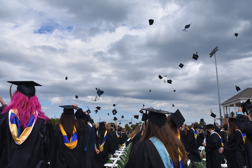 Graduates in caps and gowns celebrate outdoors by tossing their caps into the air under a cloudy sky.