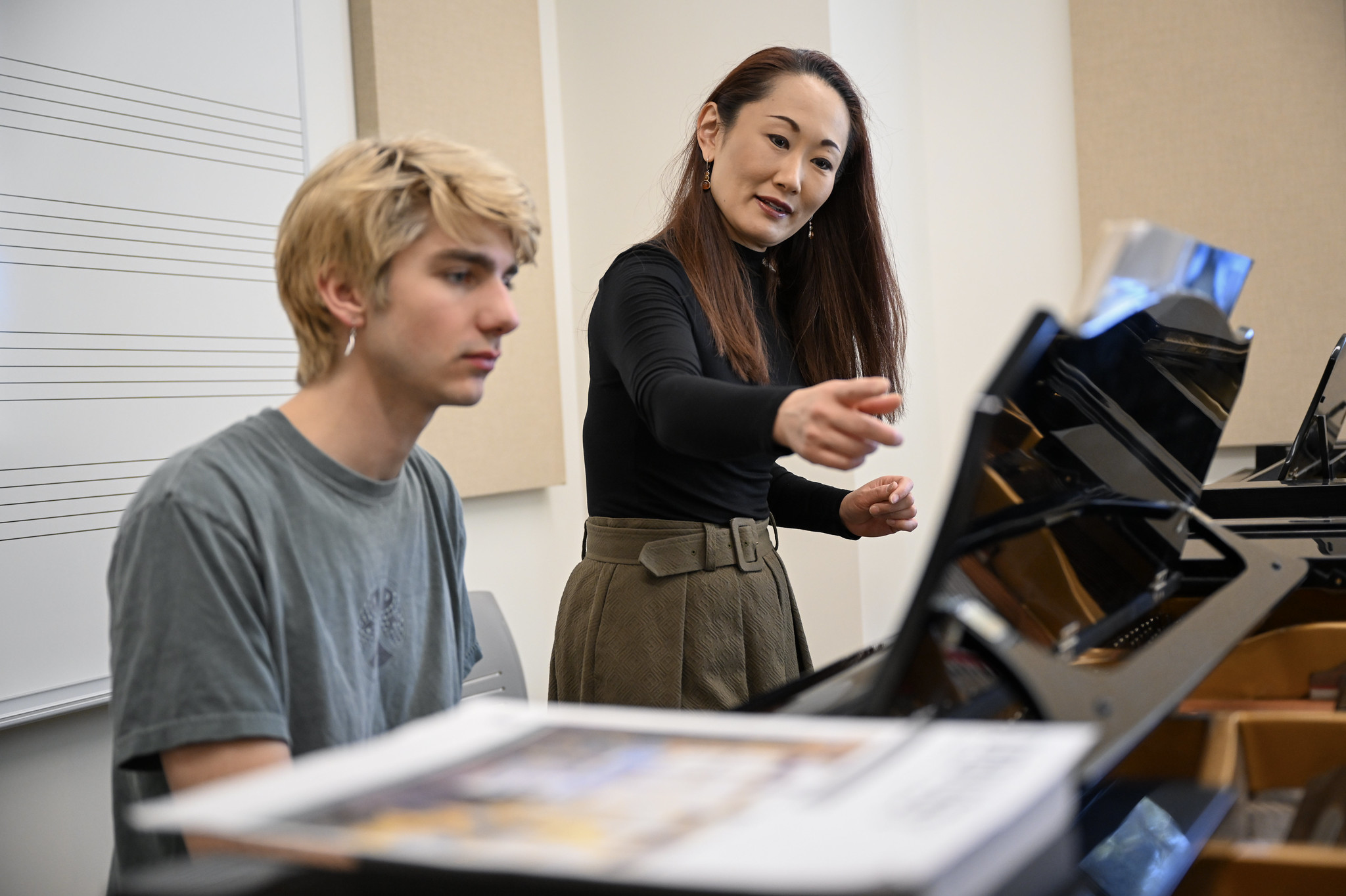 A woman instructs a young man at a piano, pointing at sheet music while he listens attentively in a music classroom.