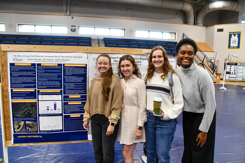 Four young women stand in front of a scientific research poster displayed in a gymnasium setting, posing and smiling for the camera.