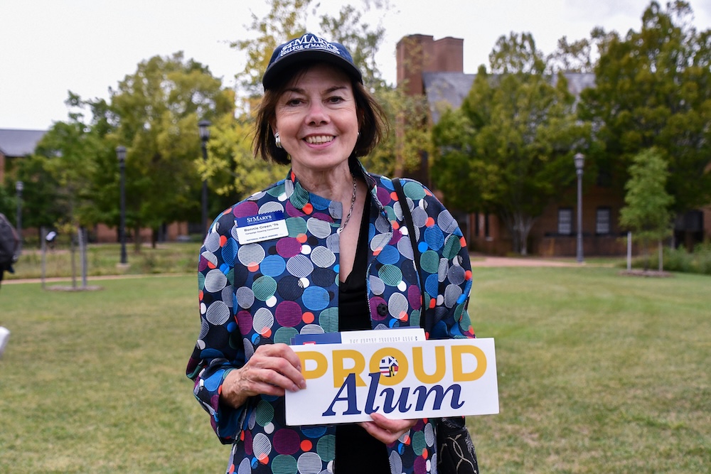 A woman wearing a patterned jacket and cap stands outdoors holding a "Proud Alum" sign, smiling at the camera. Trees and a building are visible in the background.