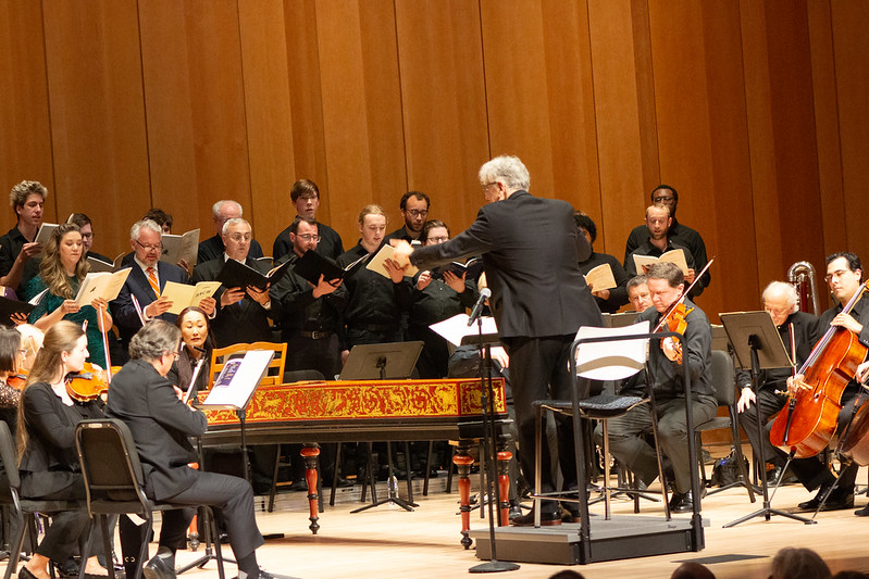 A conductor leads a choir and orchestra performing on stage. Musicians play string instruments while choir members sing, all focused on their sheet music.