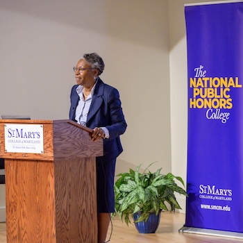A woman stands at a podium labeled St. Mary's College of Maryland, speaking, with a banner for The National Public Honors College in the background.
