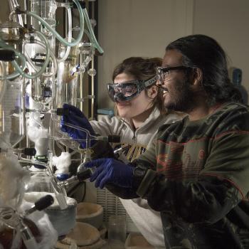 Two scientists wearing safety goggles and gloves work with glass laboratory equipment and chemical apparatus in a lab setting.