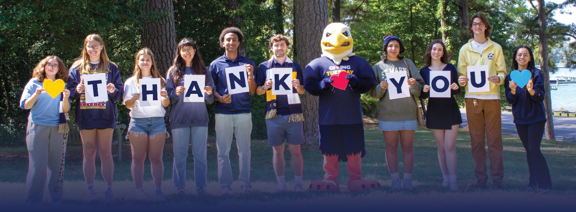 A group of people and a mascot stand outdoors holding signs that spell "THANK YOU" with hearts, on a sunny day near trees and water.