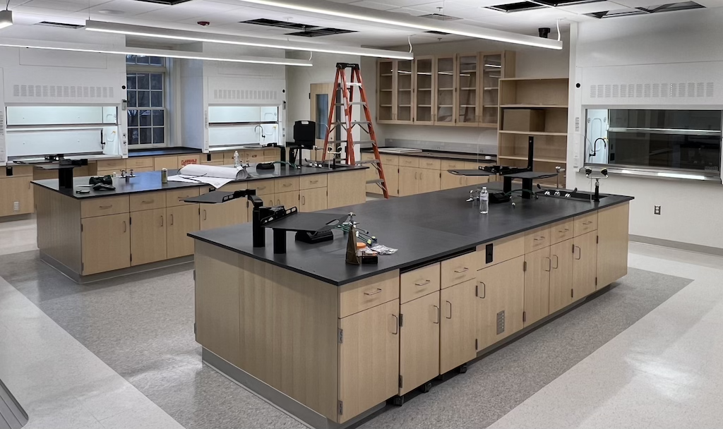 A modern science lab with wooden cabinetry, black countertops, sinks, equipment stands, a ladder, and unfinished ceiling panels.