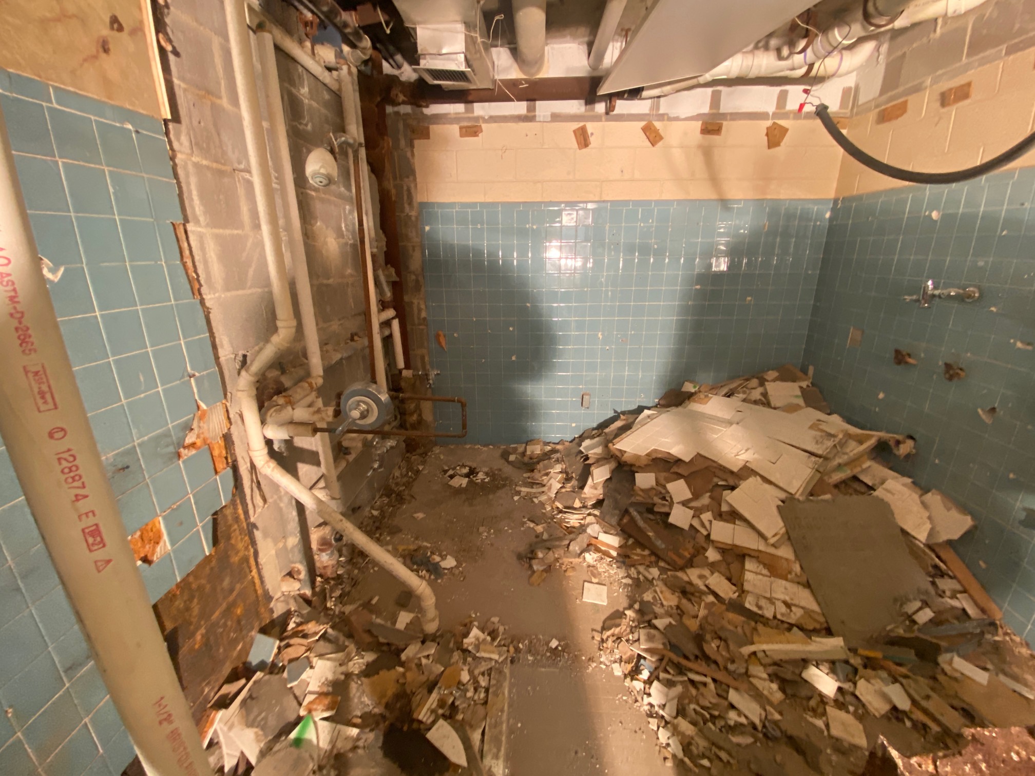 Room with damaged blue tile walls, exposed pipes, and a large pile of rubble and broken tiles covering the floor.