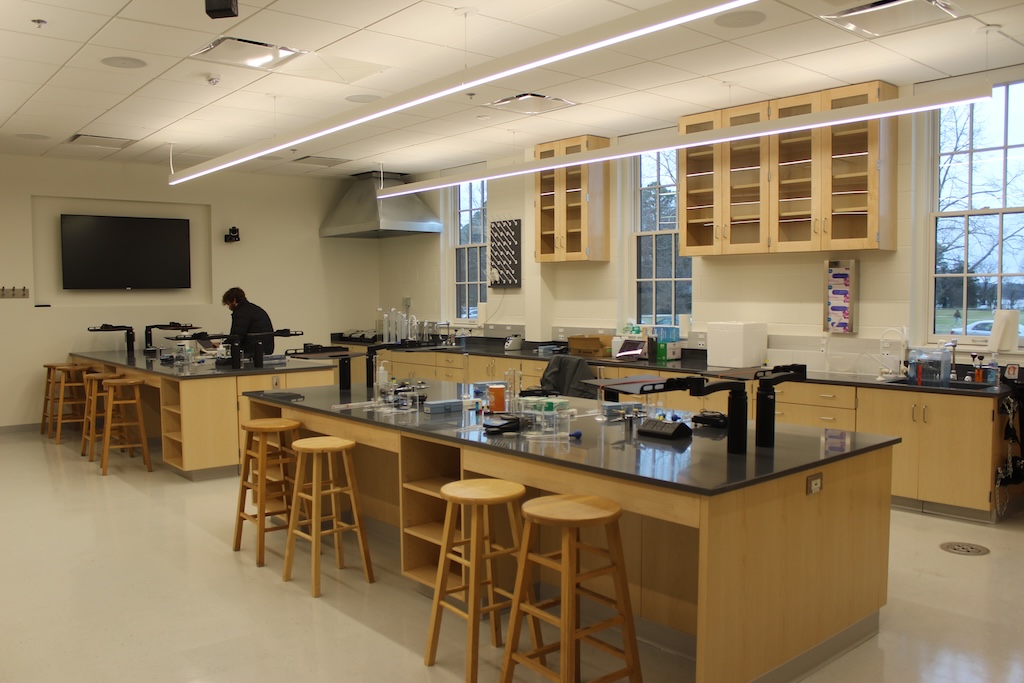 A science laboratory with wooden tables, stools, cabinets, and lab equipment. One person is working at a station. Large windows allow natural light into the room.