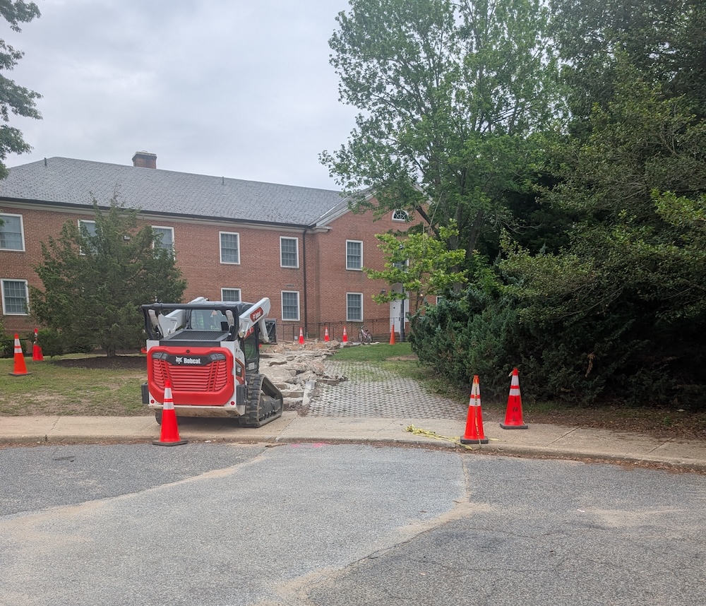 A small Bobcat loader sits on a sidewalk surrounded by orange cones and debris, next to a red brick building and trees.