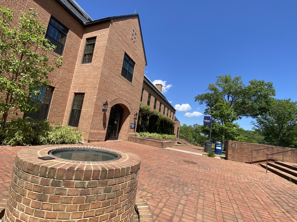 A brick building with arched entrances and a brick courtyard, featuring a round brick structure in the foreground, under a clear blue sky.