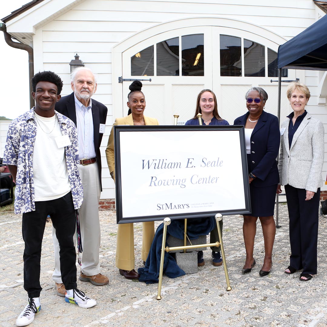 Six people stand around a large sign reading William E. Seale Rowing Center, St. Mary's outside a white building.