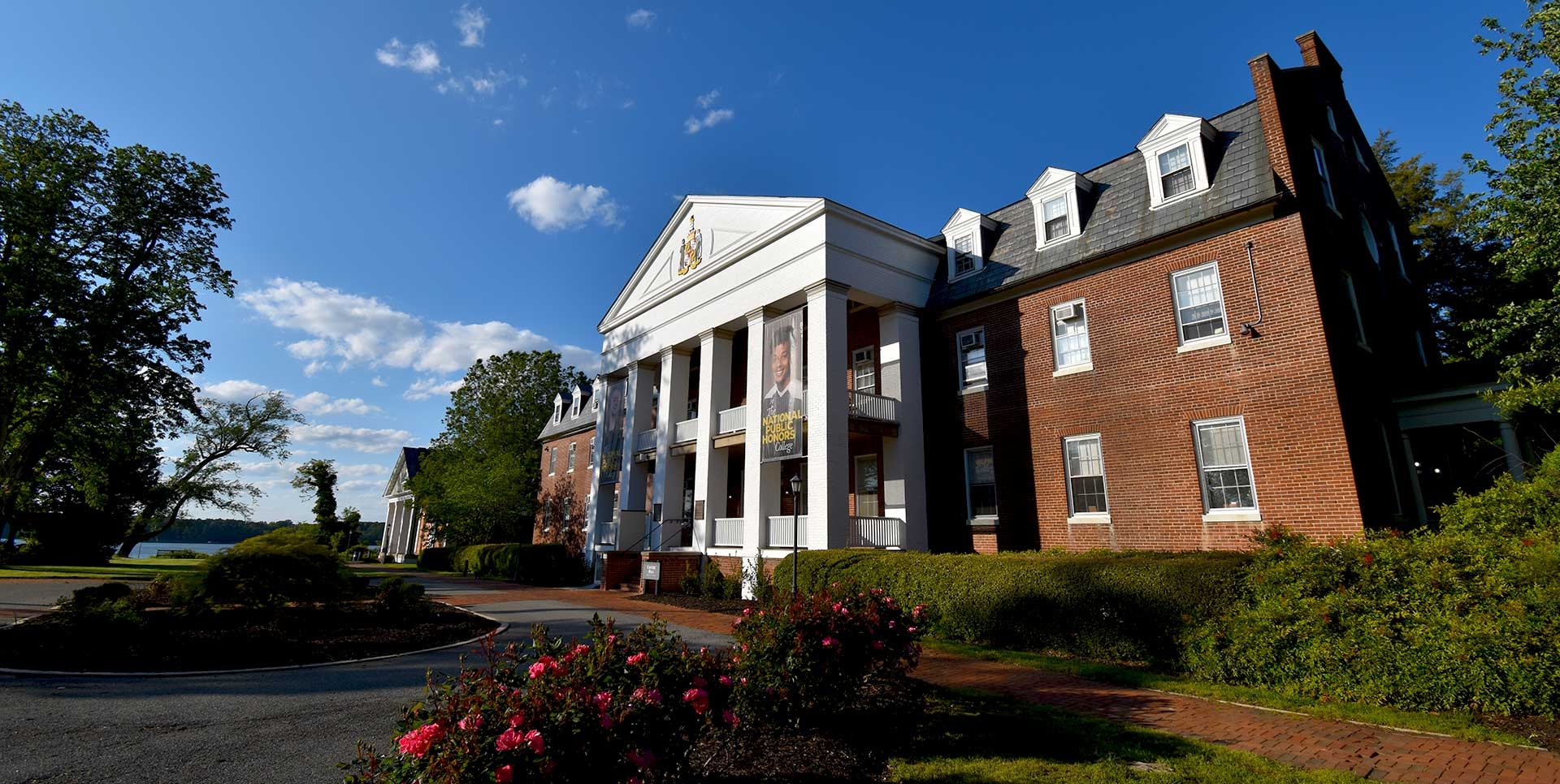 A large brick building with white columns and a triangular pediment, surrounded by bushes and flowers, under a clear blue sky.