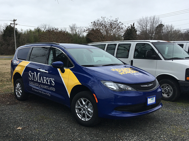 A blue minivan with "St. Mary's College of Maryland" branding is parked next to a white van in a lot on a cloudy day.