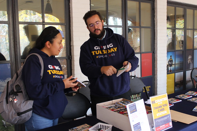 Two people wearing "Giving Tuesday" sweatshirts stand by a table with brochures and donation materials, talking at an indoor event.