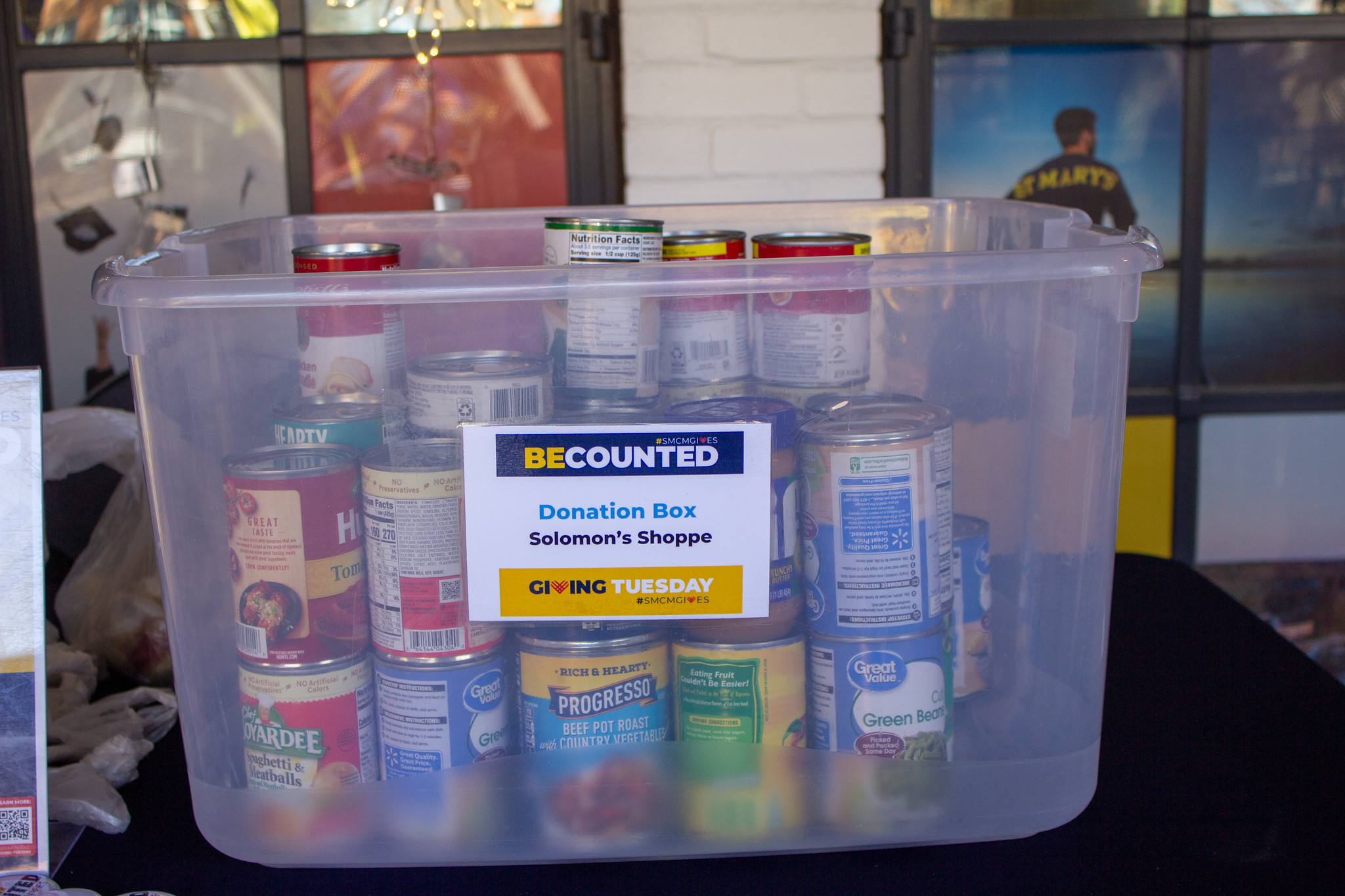A clear plastic bin filled with canned food items labeled as a donation box for Solomon’s Shoppe, promoting Giving Tuesday.