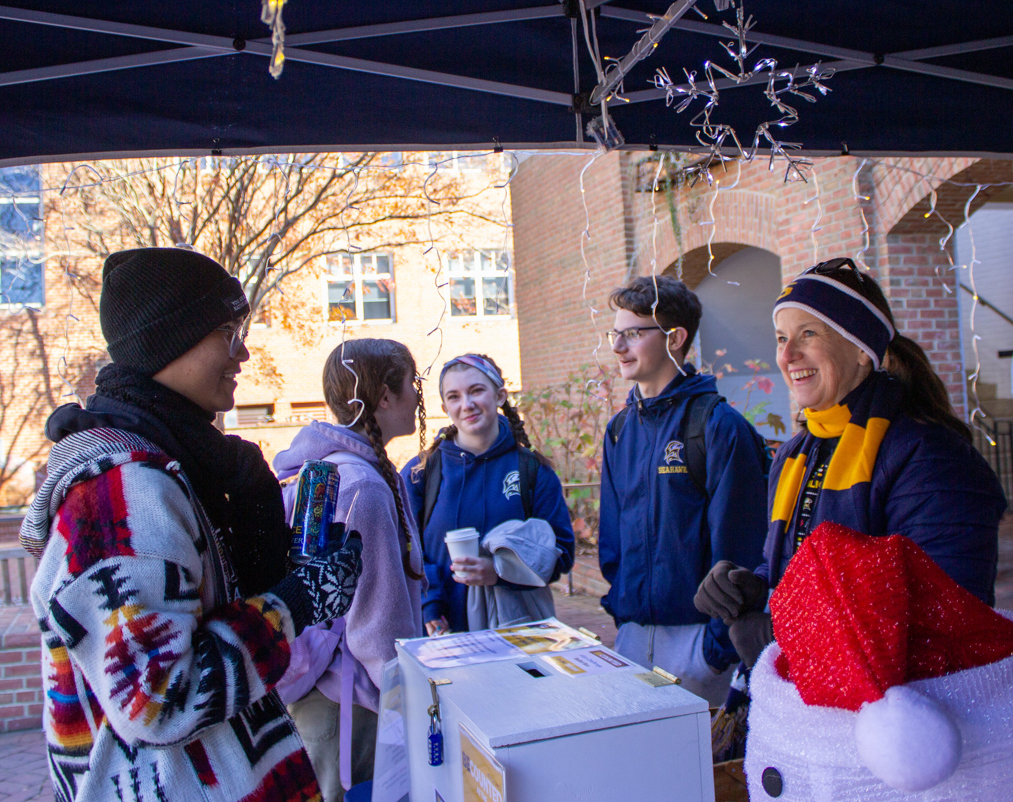 Five people dressed in winter clothing stand and converse under a canopy decorated with holiday lights, next to a table with a donation box and a Santa hat.