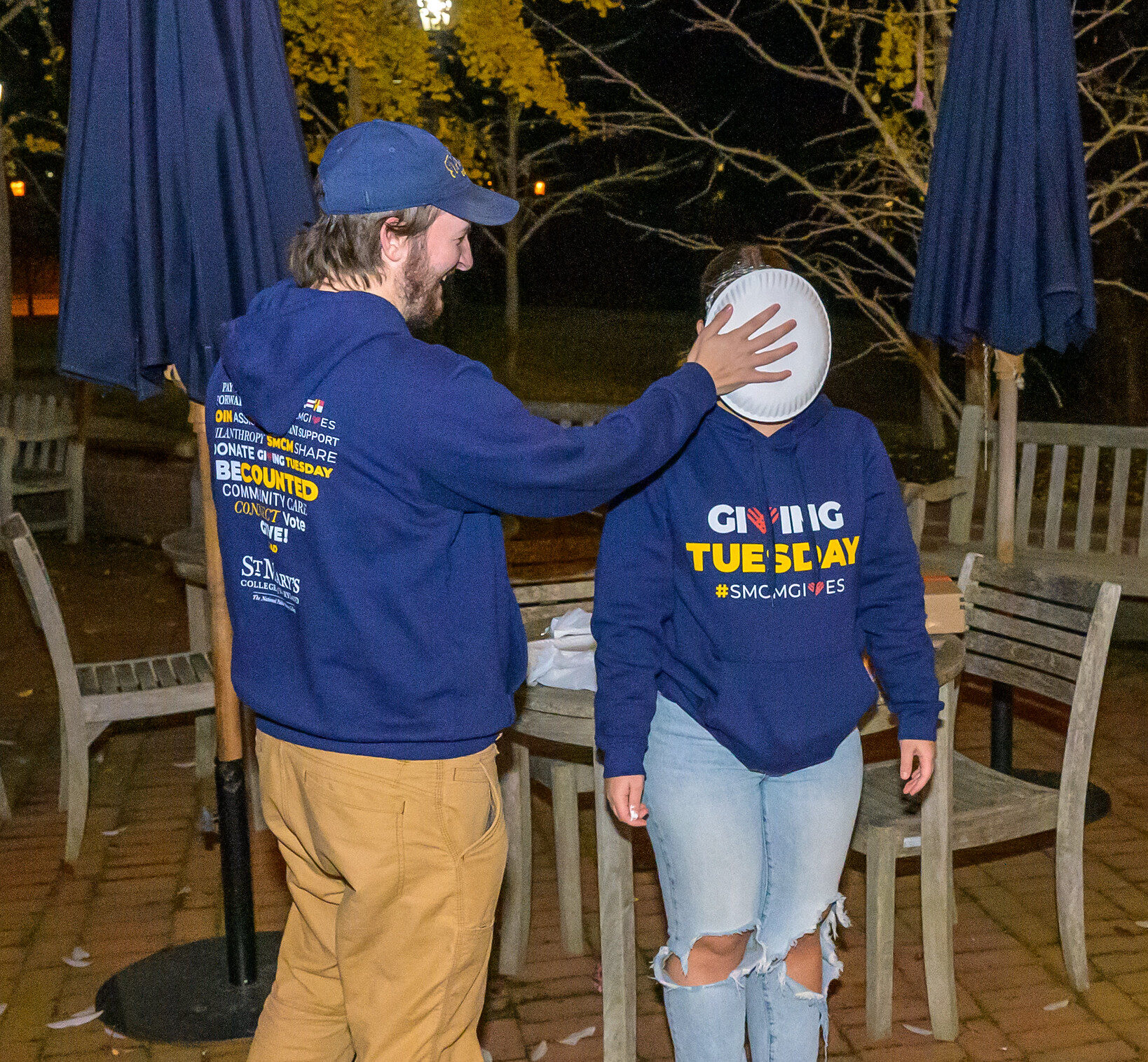 A man in casual clothes and a cap is pressing a pie into a woman's face outdoors; both are wearing "Giving Tuesday" hoodies. Broken pie plates and a sheet are on the ground.