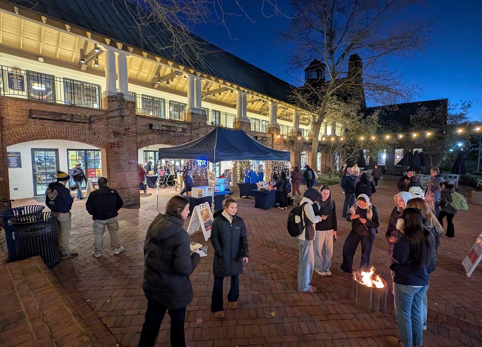 People gather in an outdoor plaza at night, some standing by a small fire, with booths and string lights set up under a covered walkway.
