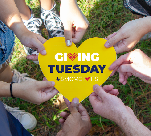 GT_5 Six people hold a yellow heart-shaped sign that reads "Giving Tuesday #SMCMGIVES" while standing on grass.
