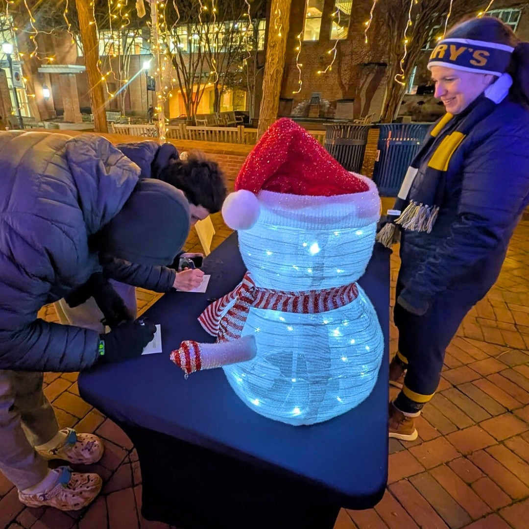 Two people at an outdoor table with a light-up snowman decoration; one is writing, while the other stands nearby wearing a winter hat and scarf.