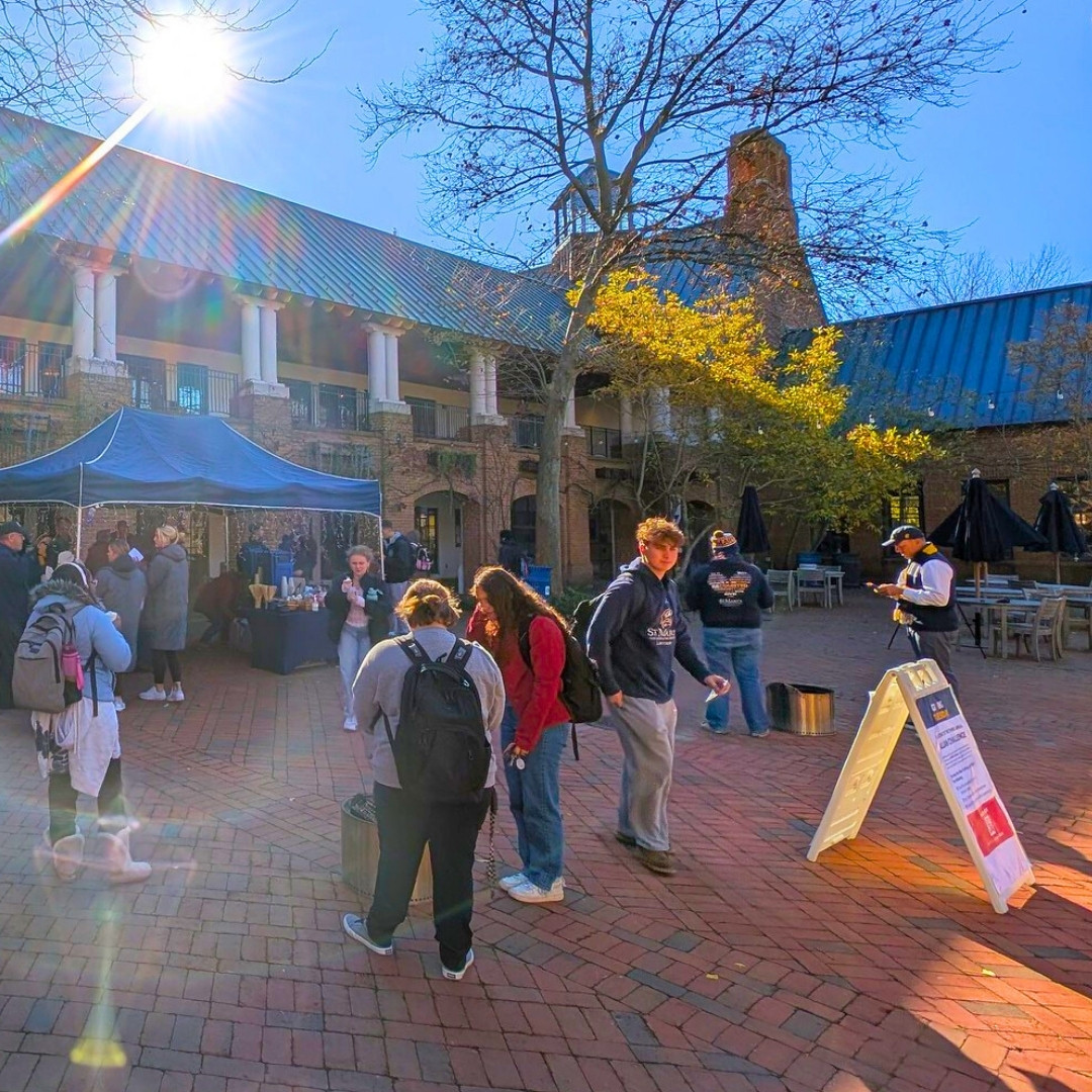 People gather in a sunny brick courtyard near a tent and informational signs, with several trees and a building in the background.